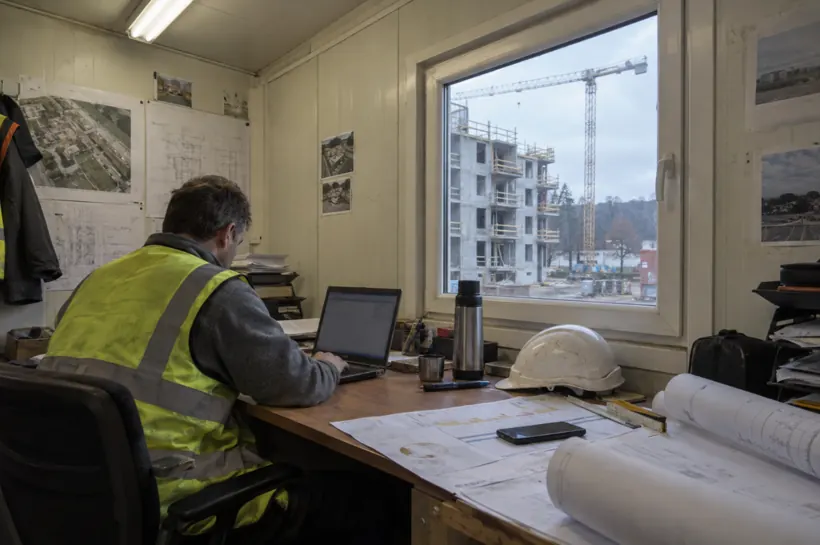 Stavbyvedoucí ve stavební buňce u notebooku, za oknem rozestavěný bytový dům a jeřáb